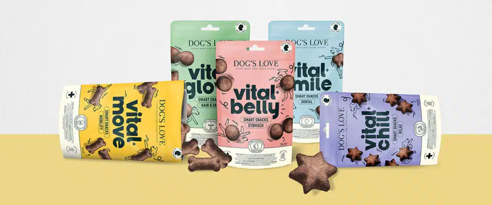 Das Bild zeigt eine Reihe von Hundesnacks der Marke „Dog's Love“ mit dem Namen „Vital Snacks“. Fünf verschiedene Verpackungen in Pastellfarben präsentieren die Sorten „Vital Move“ (Gelb, für Mobilität), „Vital Glow“ (Grün, für Fell und Haut), „Vital Belly“ (Rosa, für Magen und Darm), „Vital Smile“ (Blau, für Zahnpflege), und „Vital Chill“ (Lila, für Entspannung). Jede Verpackung ist mit Illustrationen und einem minimalistischen Design versehen, während kleine Knochen- und sternförmige Snacks aus den Verpackungen heraus zu sehen sind. Der Hintergrund ist in einem warmen Gelb- und Weißton gehalten.